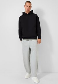 Giovane indossante un hoodie nero, pantaloni sportivi grigi e sneakers bianche, in piedi contro uno sfondo uniforme di grigio chiaro.