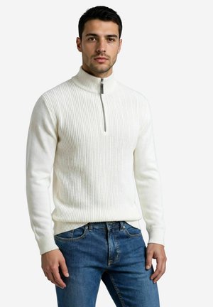 Jeune homme portant un pull blanc à demi-fermeture éclair côtelé et un jean bleu, debout devant un fond clair uni.