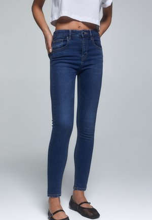 Jeans slim bleu foncé portés par une personne avec un top court blanc et des chaussures Mary Jane noires, debout devant un fond gris uni.