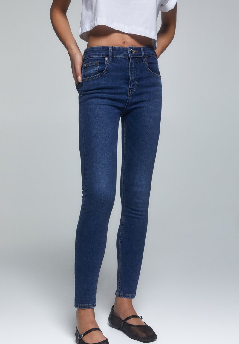 Jeans slim bleu foncé portés par une personne avec un top court blanc et des chaussures Mary Jane noires, debout devant un fond gris uni.