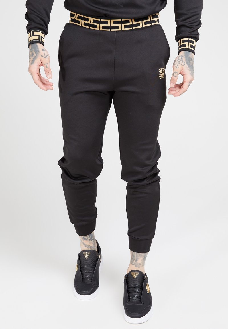 Svarta sweatpants med ett guldgeometriskt mönster på midjan, med en slim fit och ribbade manschetter. Svarta sneakers med guldaccenter.