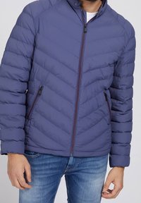 Veste matelassée bleu marine avec un motif en chevron, fermeture éclair à l'avant et poches sur les côtés. Dotée d'un col montant et de poignets côtelés pour plus de chaleur.