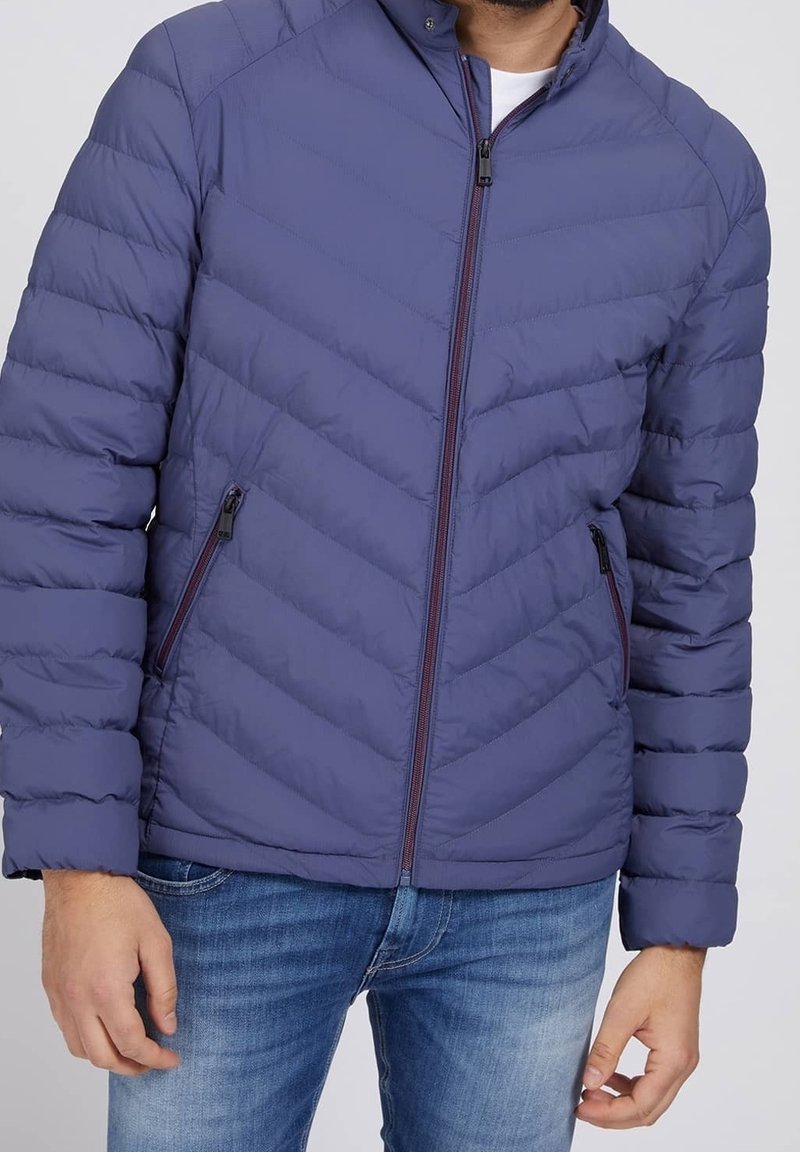 Veste matelassée bleu marine avec un motif en chevron, fermeture éclair à l'avant et poches sur les côtés. Dotée d'un col montant et de poignets côtelés pour plus de chaleur.