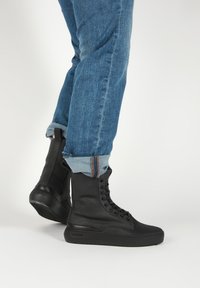 Personne portant un jean bleu retroussé et des bottes montantes noires à lacets, debout sur une surface blanche.