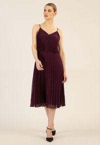 Burgunderfarbenes, plissiertes Kleid mit Spaghetti-Trägern, geschichteter Optik und fließendem Rock; kombiniert mit schwarzen Riemchen-Heels.