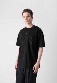 Černé tričko s krátkým rukávem vyrobené z měkkého materiálu, se zipovou kapsou na hrudi a jemným logem na přední části, volnější střih.