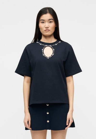 Femme portant un haut noir à manches courtes avec un décolleté en forme de goutte orné de bijoux et une jupe noire à boutons sur un fond uni.