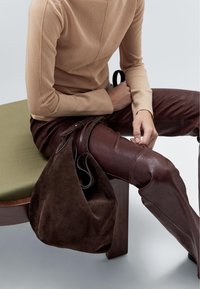 Brun suedette hobo taske med buet top, læderremme, slidt, afslappet form. Modellen bærer en beige top og brune læderbukser, siddende på en grøn pude.