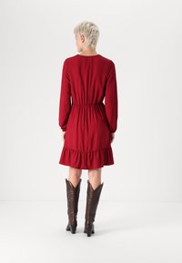 Robe rouge à manches longues avec une taille froncée et un ourlet à volants. Associée à des bottes de cowboy marron, avec des bouts pointus et une couture décorative.