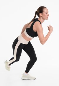 Fekete és bézs sportmelltartó és leggings szett; a leggings oldalsó fehér csíkkal rendelkezik; a modell világos színű sportcipőt visel, futó pózban áll.