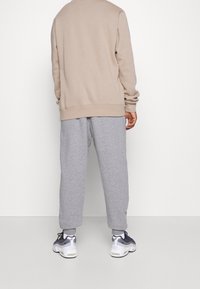 Person, der einen beigen Kapuzenpullover, graue Jogginghosen und weiße Sneakers mit blauen und schwarzen Details trägt, steht vor einer weißen Wand.
