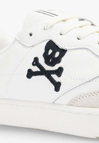 Zapatilla blanca con parte superior de cuero, que presenta un diseño bordado en negro de calavera y huesos cruzados. Acentos de ante en la puntera y el costado.