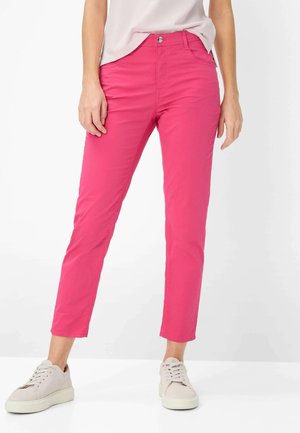 Kvinde iført knaldpink bukser med lige ben, hvide sneakers og en lyserød top, stående mod en ensfarvet hvid baggrund.