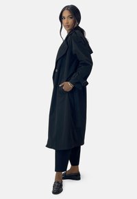 Largo abrigo negro de trench con un gran cuello, bolsillos laterales y cierre de botones. Llevado sobre pantalones negros y zapatos negros adornados.