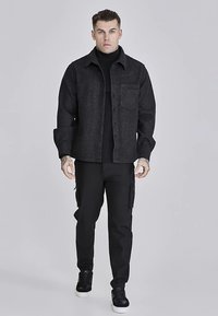 Camicia giacca in misto lana nera con colletto appuntito, chiusura con bottoni e taschino sul petto con motivo in rilievo, abbinata a pantaloni cargo neri.