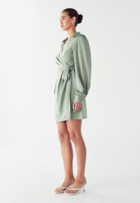 Krátkozápästové šaty v konskej zelenej farbe, vyrobené z hladkej tkaniny. Obsahujú pásik s viazaním, výstrih do V a nariasené balónové rukávy. Kombinované s priesvitnými podpätkami.