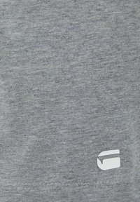 Nahaufnahme eines grauen Stoffes mit feiner Textur und einem kleinen weißen geometrischen Logo nahe der unteren Kante.