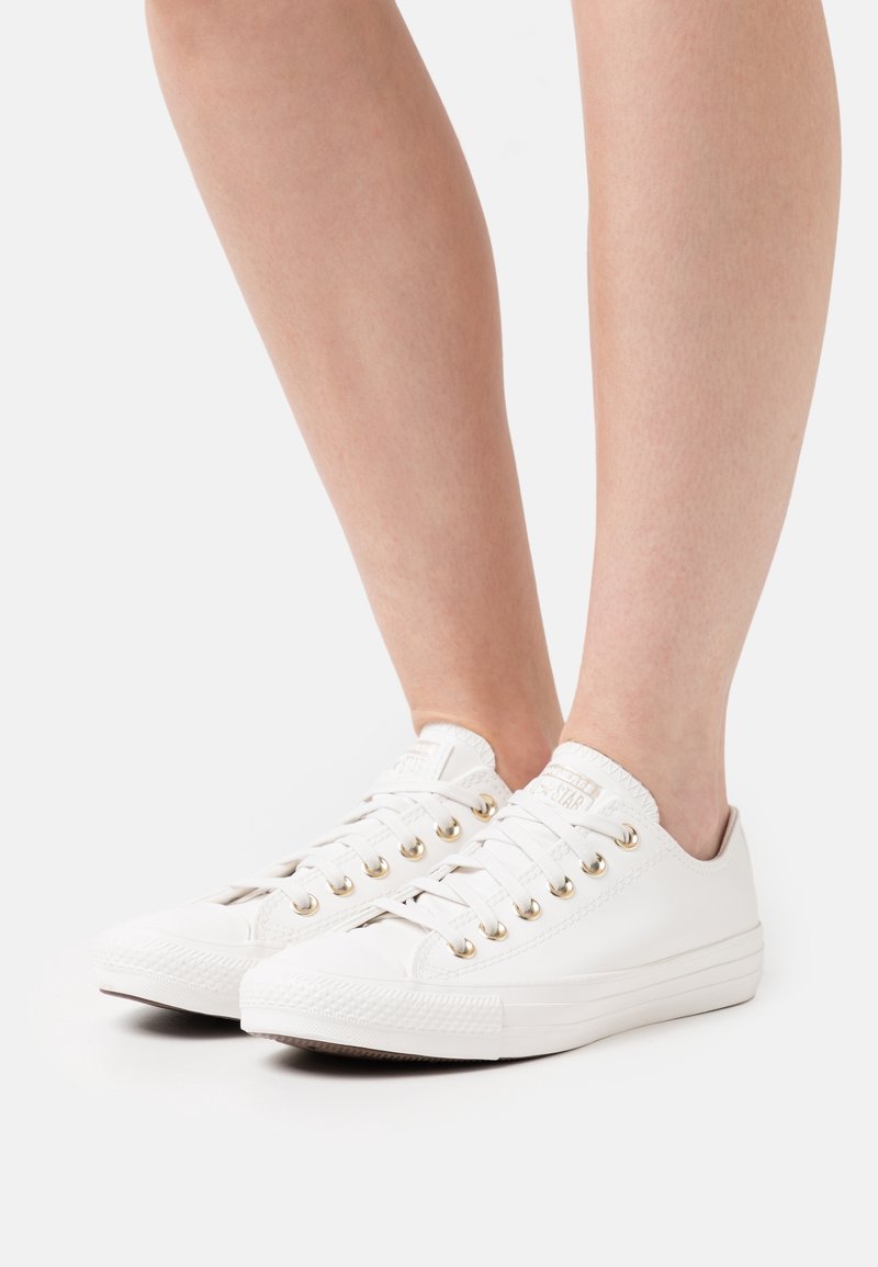 Personne portant des baskets blanches basses à œillets dorés, debout sur une surface claire, jambes nues visibles à partir du milieu du mollet.