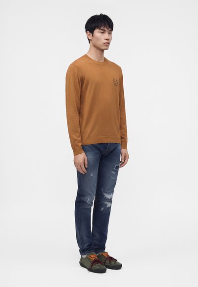 Jeune homme debout portant un pull marron, un jean bleu déchiré et des baskets vertes avec des lacets orange et rouges sur fond blanc.