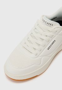 Baskets blanches avec un design perforé, lacets plats et semelle en caoutchouc. Présente un logo "SKECHERS" noir sur le côté et un talon texturé.