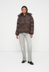 Bruine pufferjack met een hoge kraag, knoopsluiting en ritszakken. Draagt over lichtgrijze broek en zwarte schoenen. Gladde textuur.