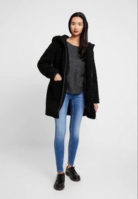 Kvinde iført sort hættefyldt fleecejakke, mørkegrå sweater, blå skinny jeans og sorte snørebåndssko, stående mod en ensfarvet baggrund.