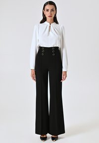 Witte blouse met een sleutelgat bij de hals, gecombineerd met hoge zwarte broeken met ronde zilveren versieringen en een brede pijpdesign.