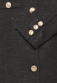 Tessuto nero strutturato con motivo intrecciato; presenta bottoni e polsini color oro. Una lucentezza notevole aggiunge profondità al design.