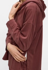 Maroon hoodie made of soft fabric with a relaxed fit, featuring a front pocket, hood, and an embroidered logo on the sleeve.