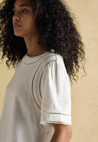 Valge puuvillane top, millel on lühikesed puff varrukad, ja detailne lõikeõmblus kaela- ja varruka servades. Pehme tekstuur.
