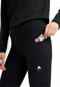 Leggings sportivi neri con una tasca laterale che contiene banconote piegate. Presentano un logo di montagna bianco sulla gamba inferiore. Texture liscia.