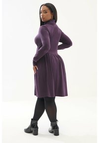 Robe longue violette à manches longues avec un col haut, un buste ajusté et une jupe évasée ; texture lisse ; associée à des bottines noires et des collants.