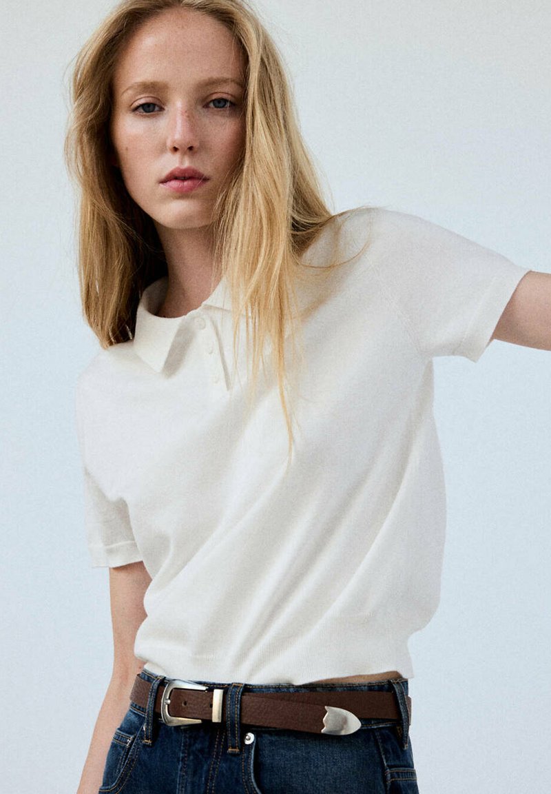 Jeune femme aux longs cheveux blonds portant un polo blanc à manches courtes rentré dans un jean bleu foncé avec une ceinture marron.