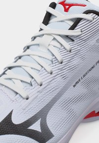 Κοντινή λήψη λευκού αθλητικού παπουτσιού με μαύρο και κόκκινο λογότυπο, λευκά κορδόνια και το κείμενο "WAVE LIGHTNING PRO" στην υφασμένη πλευρική επιφάνεια.