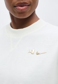 Κοντινό ενός ατόμου που φοράει λευκό πουκάμισο με χρυσά καρφίτσες με το λογότυπο της Nike στο στήθος, εμφανίζοντας μόνο τα χείλη και το κάτω μέρος του προσώπου.