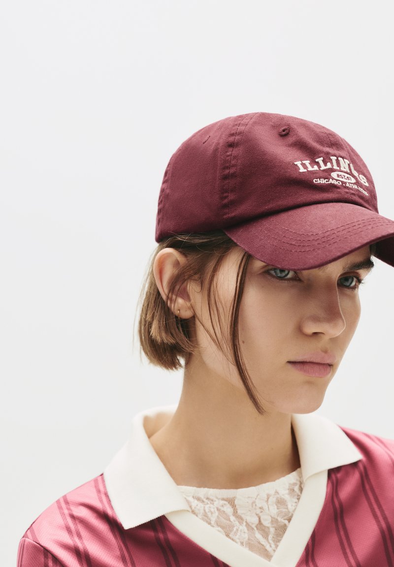 Burgundi színű baseball sapka hímzett "Illinois" felirattal. Pamut anyag, állítható pánt, rózsaszín csíkozott pólóval és fehér gallérral párosítva.