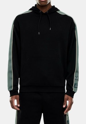 Hombre con sudadera negra con capucha y pantalones con mangas y laterales de pantalón a rayas verdes claras y blancas, con parche de logo en la manga izquierda y la pierna izquierda del pantalón.