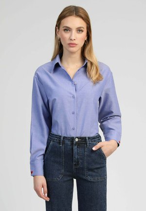 Jeune femme aux longs cheveux blonds portant une chemise bleu clair boutonnée et un jean taille haute bleu foncé, debout avec une main dans la poche.