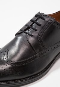 Chaussure habillée brogue en cuir noir avec design à bout ailé, détails perforés et laçage fermé sur fond blanc.