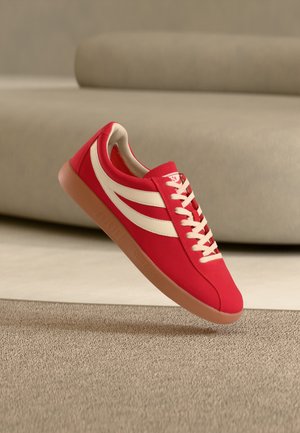 Rød joggesko med hvite striper og lisser, beige såle, svever over beige teppe med en uskarp beige sofa i bakgrunnen.