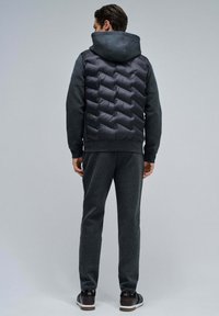 Gilet en doudoune gris avec un design texturé, associé à un pantalon de survêtement gris foncé. Le gilet dispose d'une capuche et d'un motif matelassé à l'arrière.