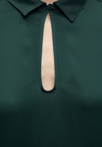 Chemise col vert foncé avec un décolleté vertical en forme de trou de serrure laissant apparaître la peau en dessous, fermé en haut par un seul bouton noir.