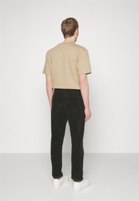 Chemise beige à manches courtes associée à un pantalon en velours côtelé noir et des baskets blanches, vue de dos sur un fond uni.