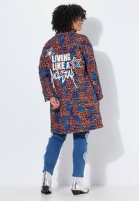 Kleurige dierenprint jas met oranje, blauwe en zwarte patronen; voorzien van witte grafische tekst op de rug; gecombineerd met versleten jeans en zilveren laarzen.