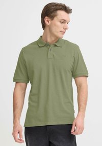 Polo vert olive en tissu texturé, avec deux boutons bleus et un petit logo sur la poitrine. Manches courtes et col classique.