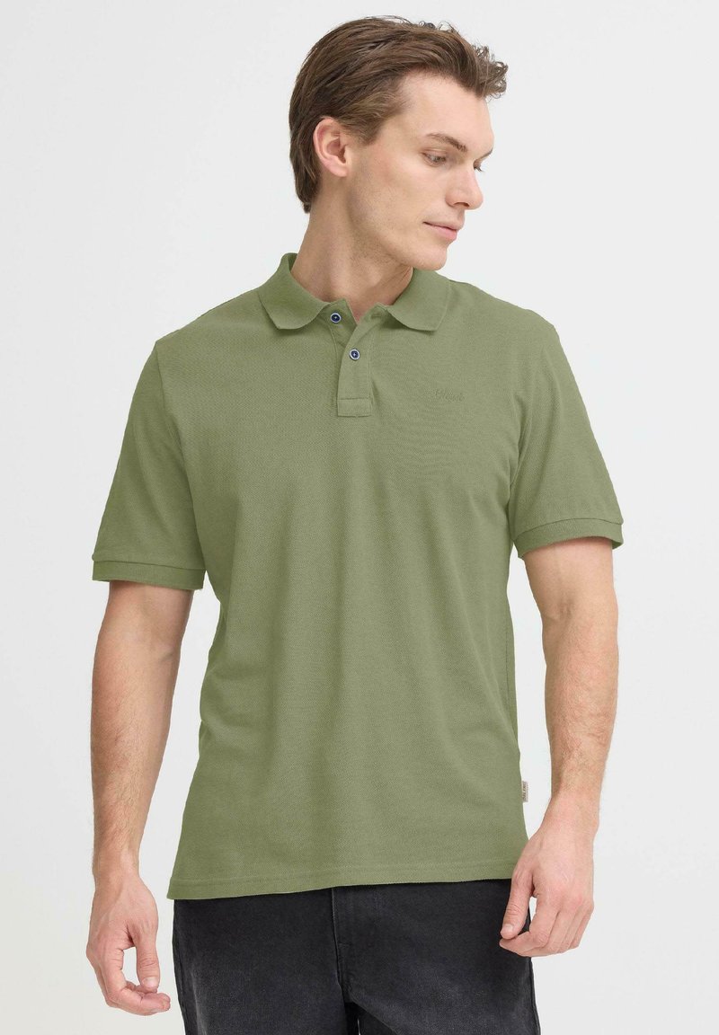 Polo vert olive en tissu texturé, avec deux boutons bleus et un petit logo sur la poitrine. Manches courtes et col classique.