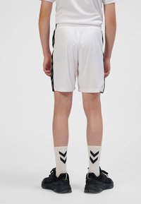 Weiße Sportshorts mit schwarzen Seitenstreifen, getragen mit cremefarbenen Socken, die ein schwarzes Chevronmuster aufweisen, und schwarzen Sportschuhen.