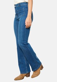Jeans de mezclilla azul de cintura alta con un diseño de pierna acampanada, que presentan dos bolsillos traseros y un desvanecimiento mínimo. Combinados con botas de tacón marrones.