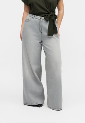 Žena má na sebe svetlosivé široké rifle s vysokým pásom, v kombinácii s tmavozelenou krátkym rukávom, na ktorý je previazaná blúzka, vidieť čierne topánky.