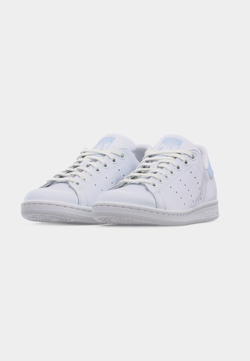 Par af hvide lavtop-sneakers med lyseblå hælkiler, perforerede sidetaljer og hvide snørebånd på en ensfarvet baggrund.